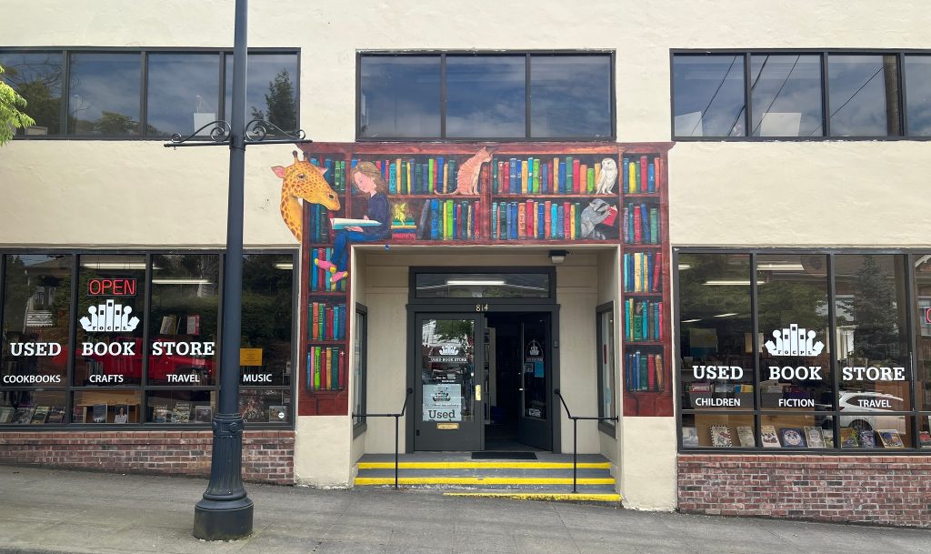 Friends of Oregon City Library Used Book Store mural - bookshelves  with a girl sitting on one of the shelves reading a book and some animals framing the door of the bookstore